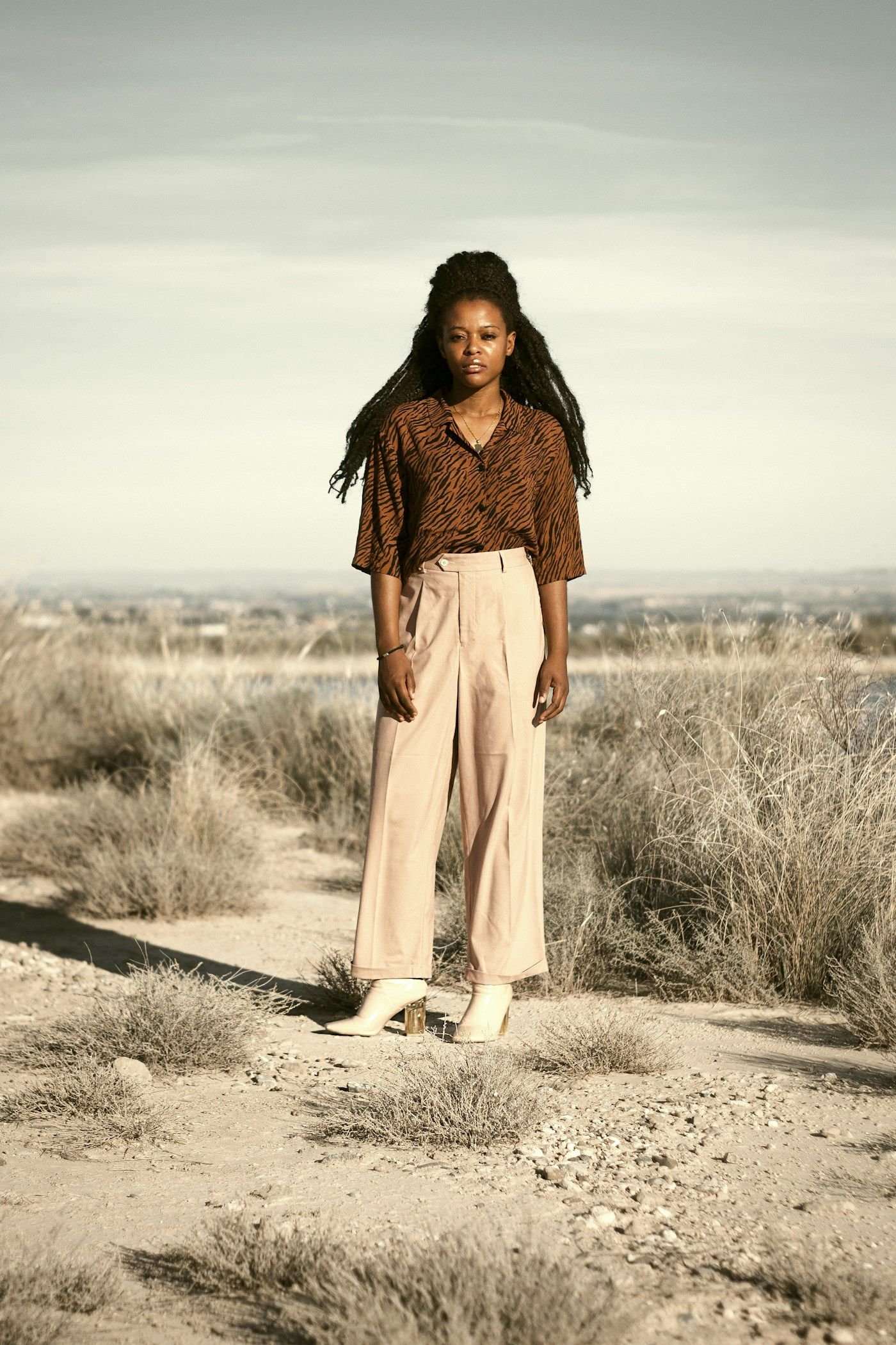 Model in cream wide-leg silk trousers and a soft blouse standing in open dry grassland