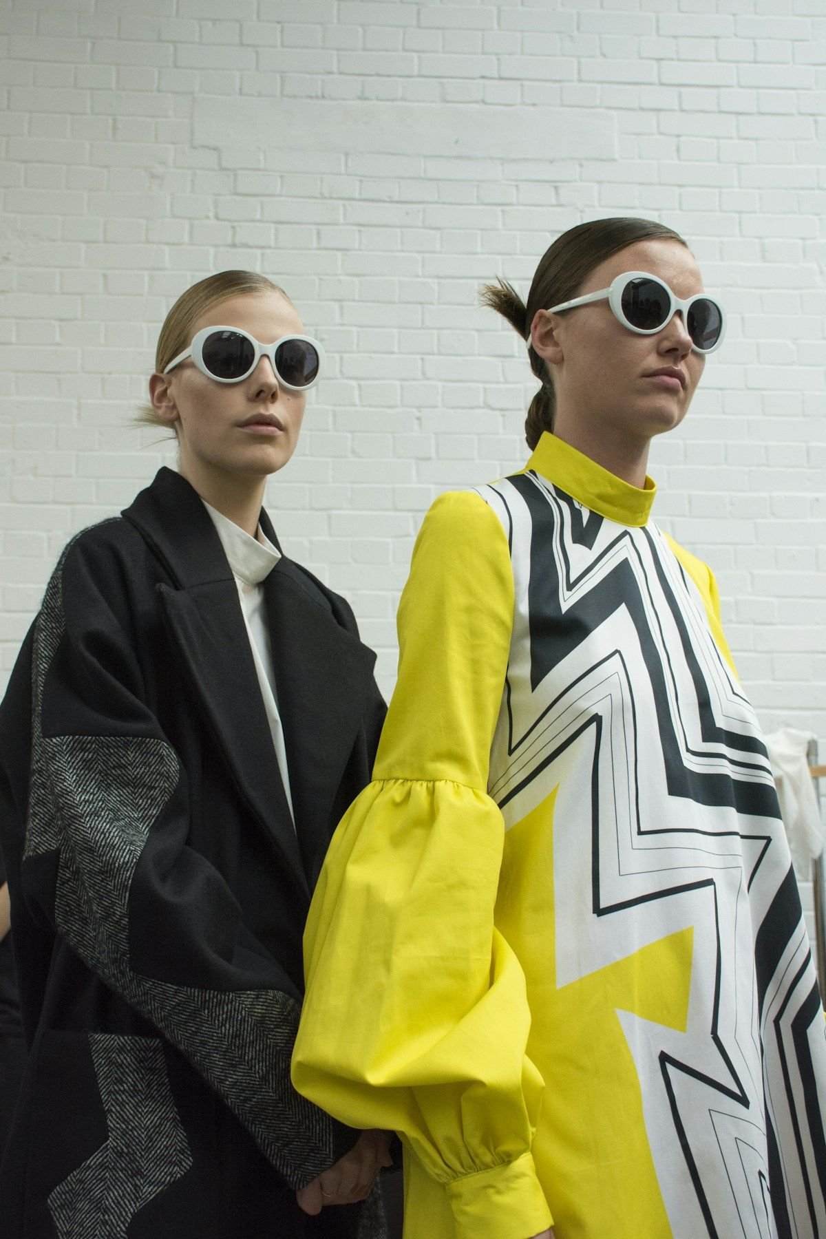 Two models in bold graphic outerwear standing side by side before a white brick wall