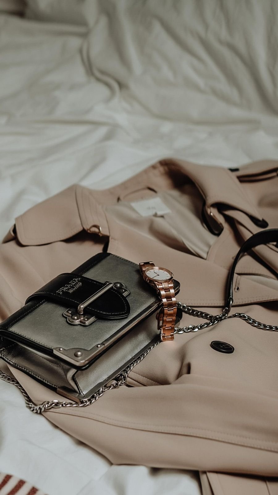 Camel trench coat and a small structured handbag arranged on pale bedlinen