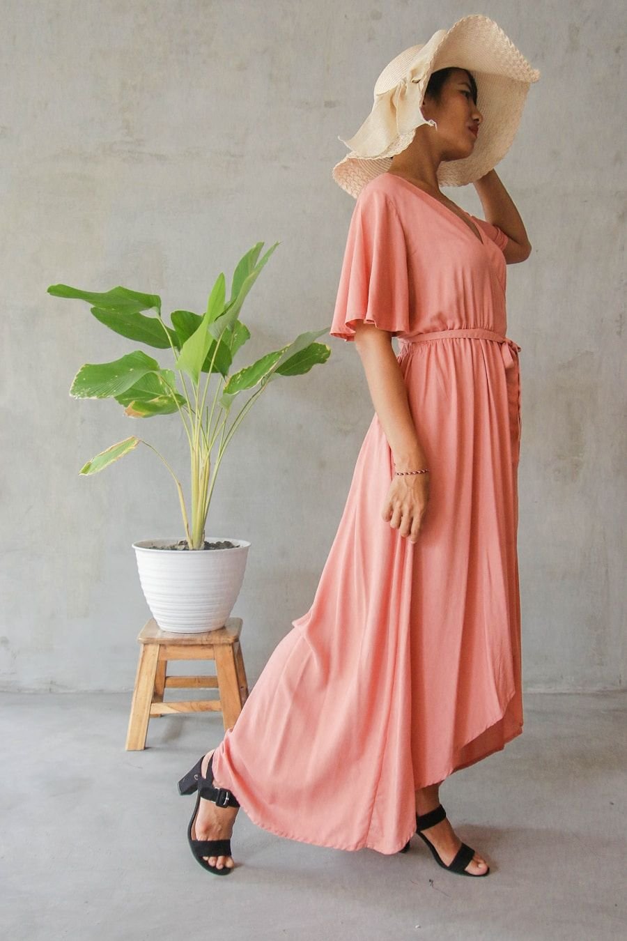 Model in a soft peach silk column dress and wide-brimmed straw hat beside a green plant